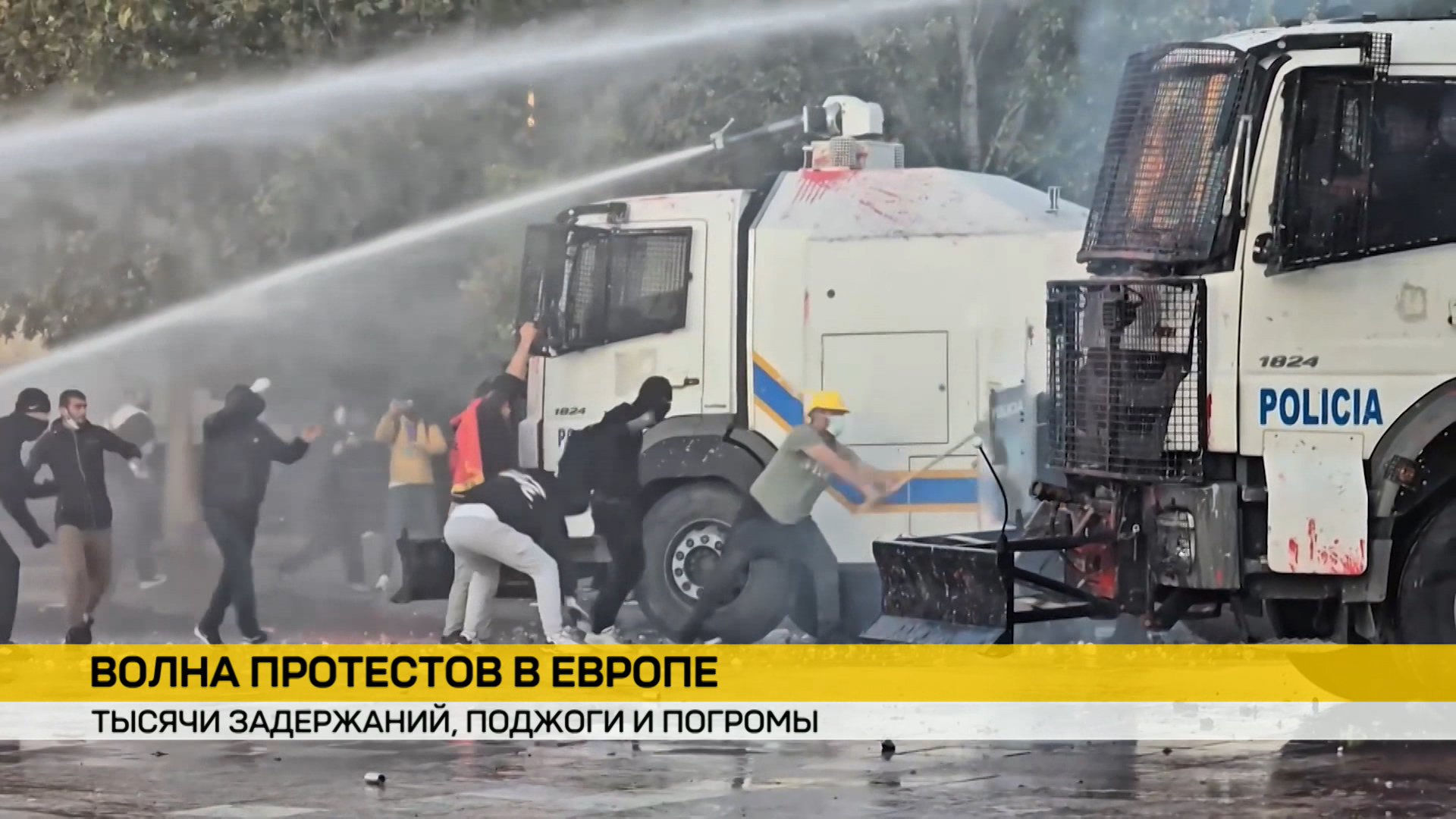 Протестующие в масках атакуют полицейские грузовики с надписью «POLICIA». Спецтехника применяет водометы для разгона толпы среди дыма.